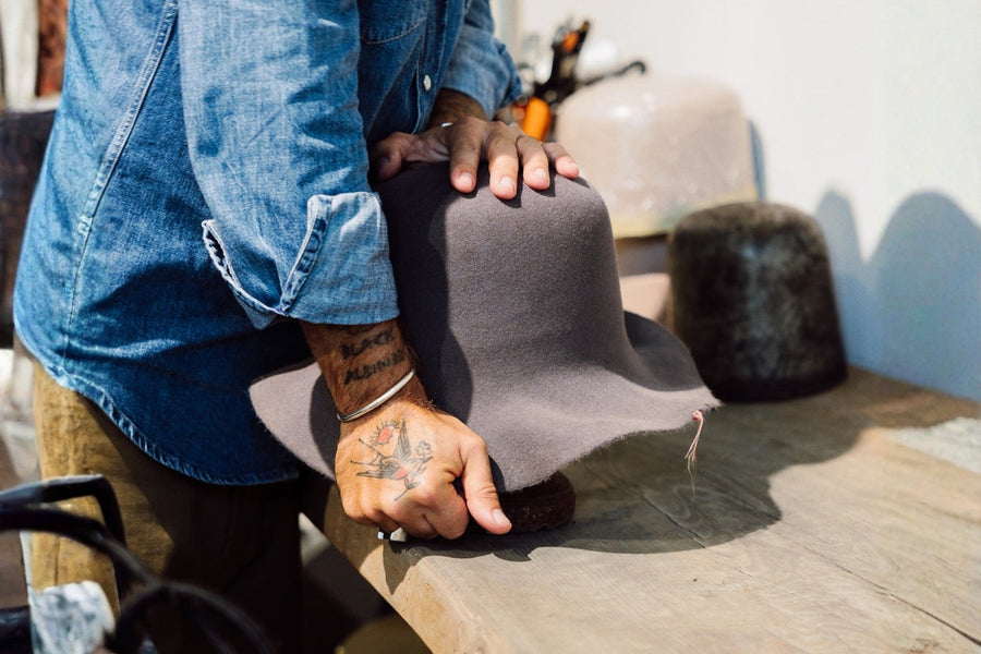 Craftsman shaping grey felt hat by hand on wooden table in a workshop