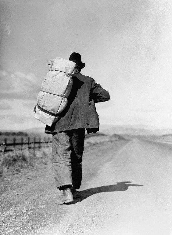 Man in vintage hat and suit walking on dirt road with large backpack, black and white photo