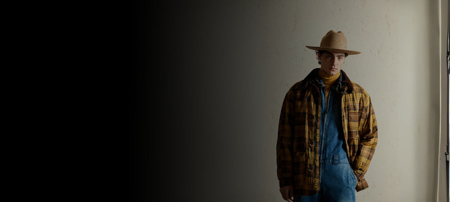 Man in wide-brimmed felt hat, yellow plaid jacket, and denim overalls by a wall